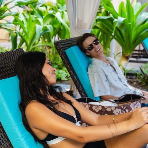 Couple relaxing on lounge chairs inside shaded pool cabana at Hilton Hawaiian Village with tropical greenery