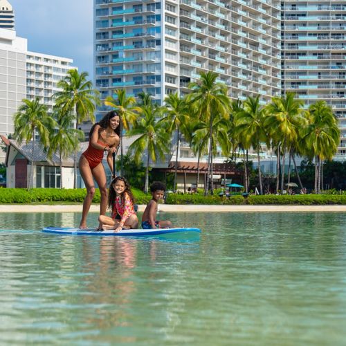 Mother and children enjoying stand-up paddle boarding on calm Waikiki lagoon with resort towers behind