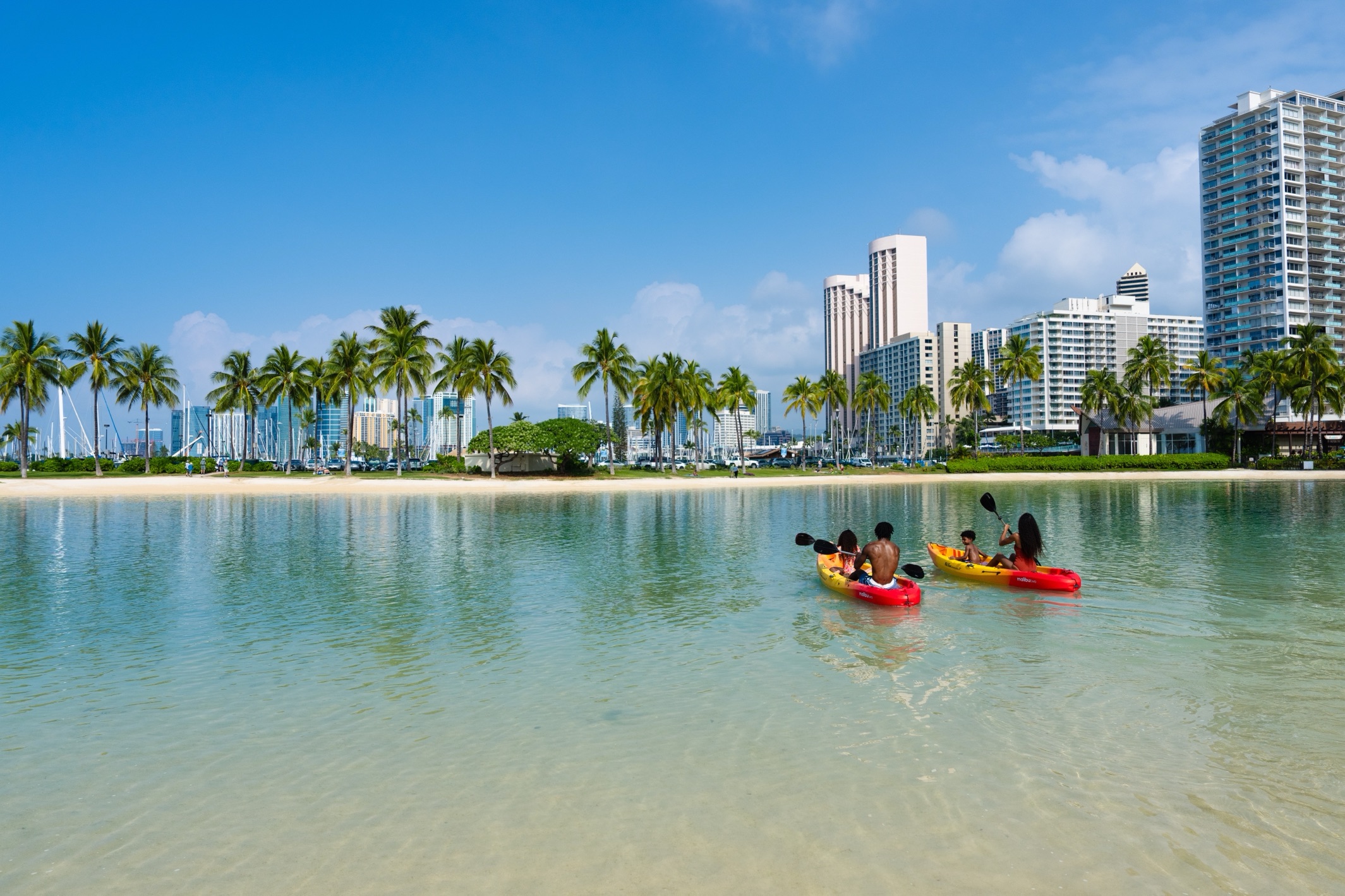 Family kayaking together in colorful kayaks on calm Waikiki lagoon with palm trees and resort beach