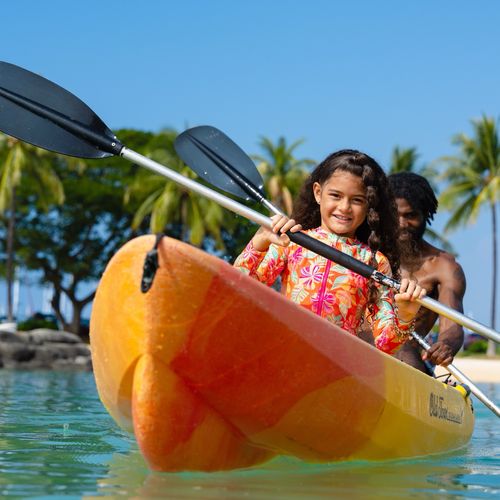 Young girl kayaking in orange kayak with adult guide in tropical Hawaii lagoon with palm trees