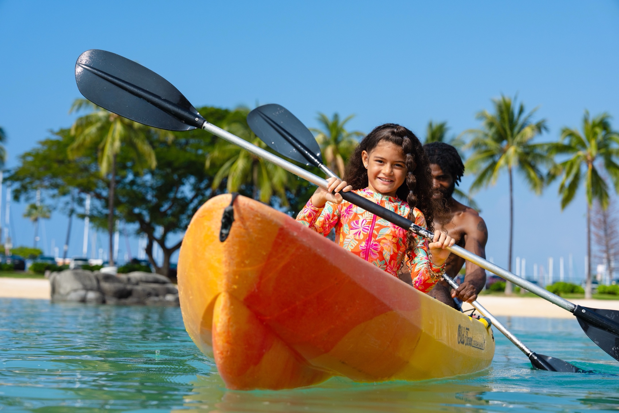 Young girl kayaking in orange kayak with adult guide in tropical Hawaii lagoon with palm trees